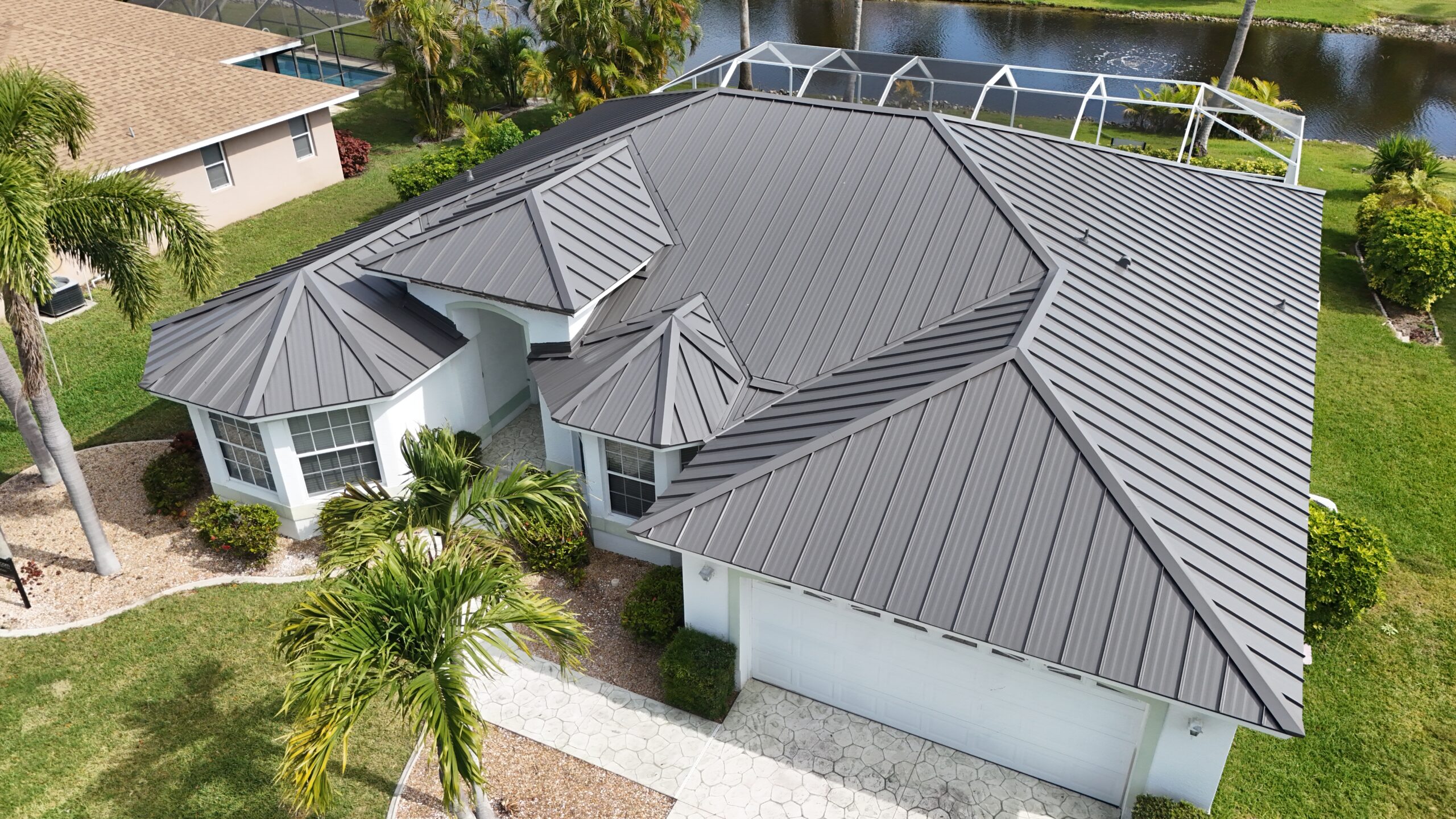 Aerial view of a home with a dark gray metal roof, surrounded by palm trees and a waterfront view.