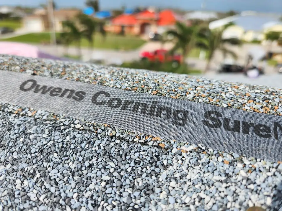 A close-up, eye-level shot of Owens Corning SureNail shingles. The SureNail technology is a wide, woven fabric strip with "Owens Corning SureNail" printed on it, embedded in a layer of small, gray, and black granules. The background is a blurry residential area with palm trees and houses under a partly cloudy sky.