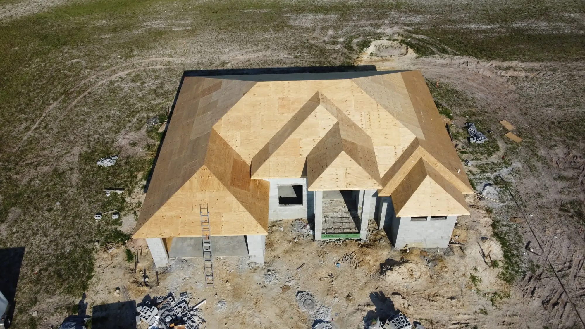 Aerial view of a new house under construction with the roof sheathing complete, surrounded by dry, undeveloped land.