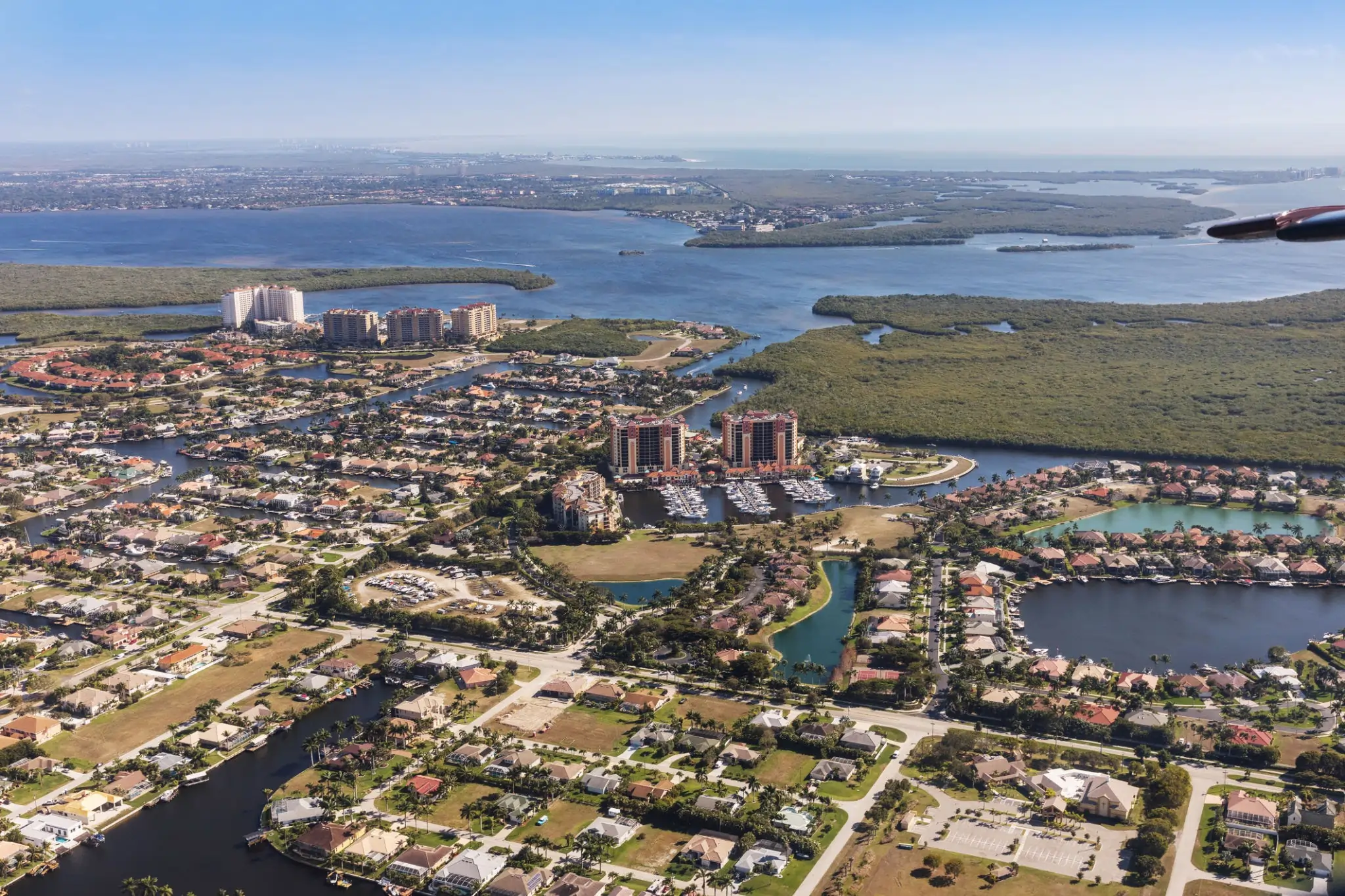 High-angle aerial view of a sprawling Florida-style coastal community with numerous waterfront homes, luxury high-rise apartment buildings, a boat-filled marina, and winding canals surrounded by green mangrove islands and a large bay in the background.