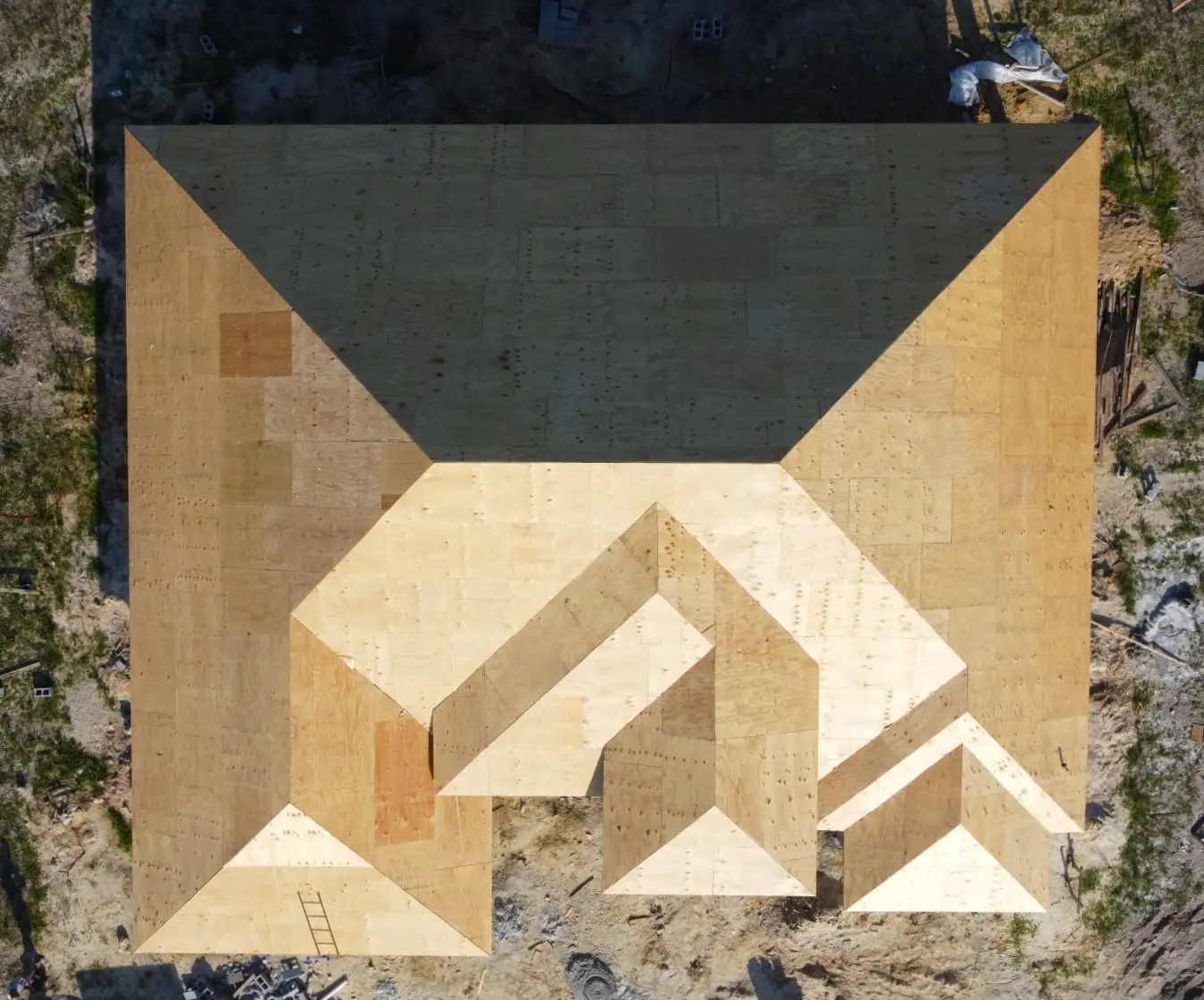 A high-angle drone shot showing the complex hip and valley roof structure of a new house, completely covered in light-colored plywood sheathing.